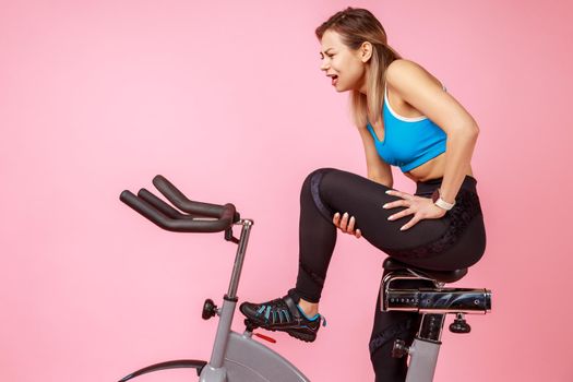 Side view portrait of athletic woman has workout at home, riding exercise bike and injured her leg, feeling pain in thigh, wearing sports tights and top. Indoor studio shot isolated on pink background