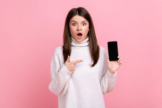 Shocked girl with open mouth pointing at empty smart phone display with copy space for advertisement, wearing white casual style sweater. Indoor studio shot isolated on pink background.