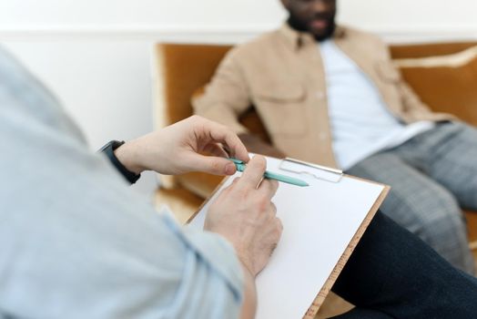 A male psychologist conducts psychological therapy with an African American man, makes notes in a clipboard