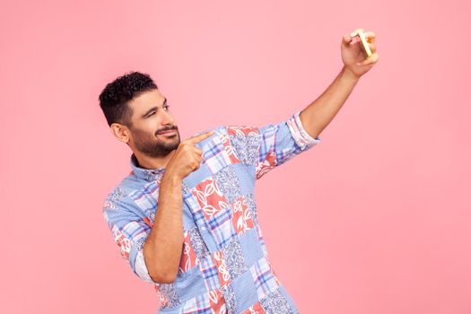 Hey you there. Friendly brunet bearded wearing blue casual style pointing finger to cellphone, smiling talking video call on mobile phone. Indoor studio shot isolated on pink background.