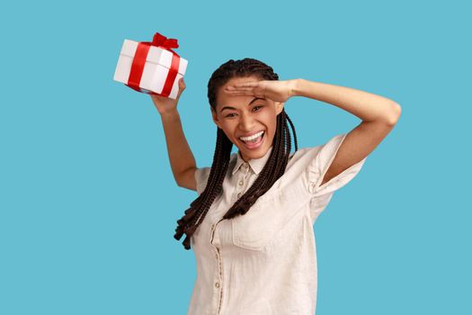 Portrait of excited satisfied woman with black dreadlocks holding wrapped present box, looking far away with hand over forehead, wearing white shirt. Indoor studio shot isolated on blue background.
