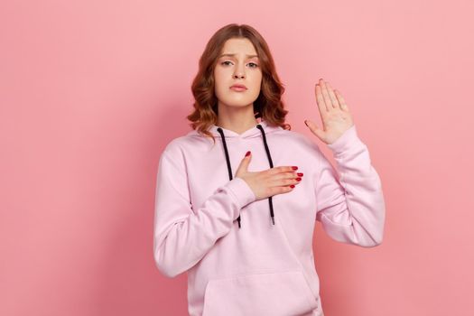 Portrait of sincere honest teen girl in hoodie holding hand up and on chest, pledging allegiance, taking oath with responsible expression. Indoor studio shot isolated on pink background