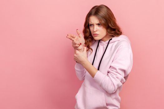 Portrait of curly-haired teen girl in hoodie pointing finger gun to camera, aiming and threatening to shoot with pistol hand gesture. Indoor studio shot isolated on pink background