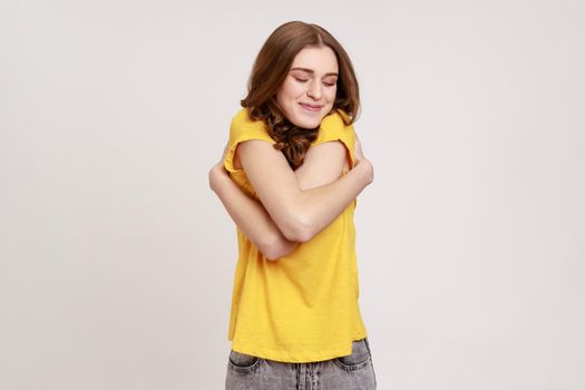 I am egoist, love myself. Selfish narcissistic teen girl with wavy hair embracing herself and smiling, closed eyes with dreamy satisfied expression. Indoor studio shot isolated on gray background.