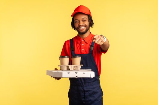 Happy smiling deliveryman wearing blue overalls holding take away coffee and pizza in cardboard box, pointing at camera, your delivery order. Indoor studio shot isolated on yellow background.