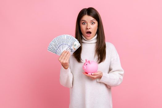 Surprised brunette female holding fan of dollar bills and piggy bank, savings, growth of deposit, wearing white casual style sweater. Indoor studio shot isolated on pink background.