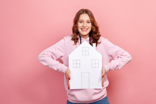 Portrait of excited curly haired teenage girl in hoodie holding paper house looking at camera with toothy smile, dreaming of own home. Indoor studio shot isolated on pink background