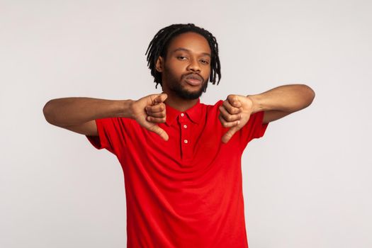 Bearded man with dreadlocks wearing red casual style T-shirt, showing thumbs down, expressing disapproval, criticizing and disagreeing with suggestion. Indoor studio shot isolated on gray background.