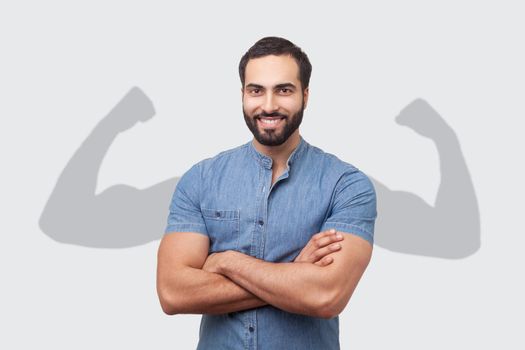 Successful positive bearded man in blue shirt folding hands on chest and looking at camera with toothy smile showing strong big arms in shadow. Indoor studio shot isolated on gray background