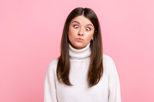 Idiot woman making silly humorous face with eyes crossed, showing comical silly brainless expression, wearing white casual style sweater. Indoor studio shot isolated on pink background.