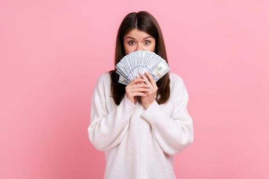 Portrait of shy female with dark hair covering half of face with dollar banknotes, looking at camera, wearing white casual style sweater. Indoor studio shot isolated on pink background.