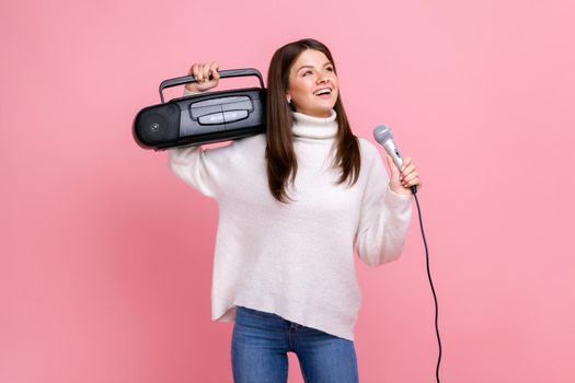 Portrait of happy positive female having fun, holding record player and singing via microphone, wearing white casual style sweater and jeans. Indoor studio shot isolated on pink background.