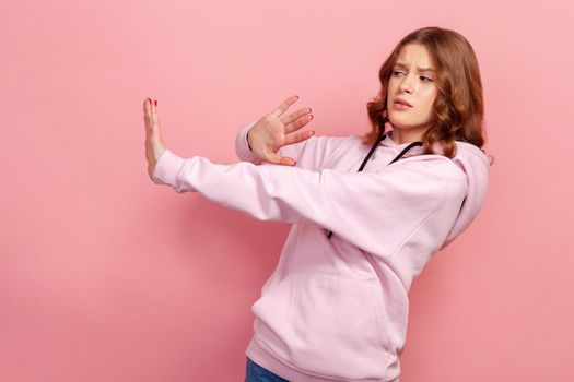 Side view of scared young girl standing with raised hands, gesturing stop, panicking looking frightened of danger, phobia. Indoor studio shot isolated on pink background