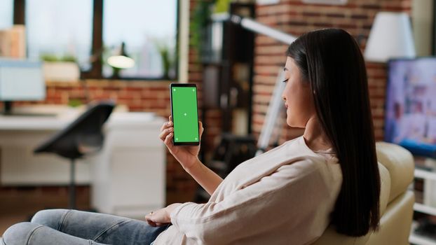 Young adult person sitting on couch with mobile cellphone having mockup chroma key background. Asian woman having smartphone with green screen display while sitting at home on sofa. Tripod shot