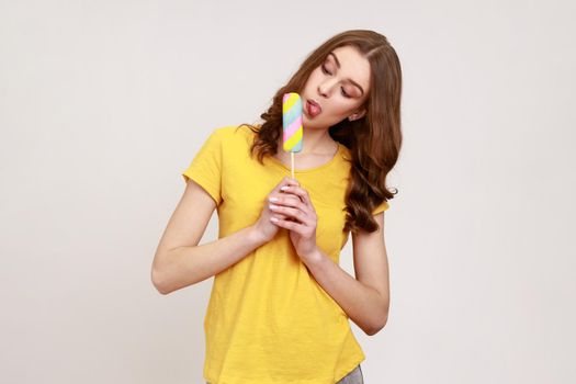 Portrait of funny teenager girl sticking out tongue licking ice cream, tasting and enjoying delicious flavor dessert, sweet confection. Indoor studio shot isolated on gray background.
