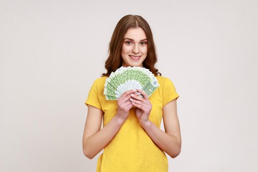 Rich beautiful woman of young age with brown wavy hair in yellow casual style T-shirt showing euro banknotes, high salary, big profit. Indoor studio shot isolated on gray background.