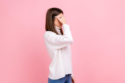 Side view portrait of brunette woman covering eyes with palm, feeling depressed of what she saw, wearing white casual style sweater. Indoor studio shot isolated on pink background.