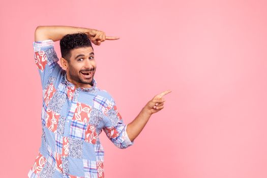 Wow, attention to advertising. Portrait of happy bearded man pointing to side copy space with both hands and showing blank wall for idea presentation. Indoor studio shot isolated on pink background.