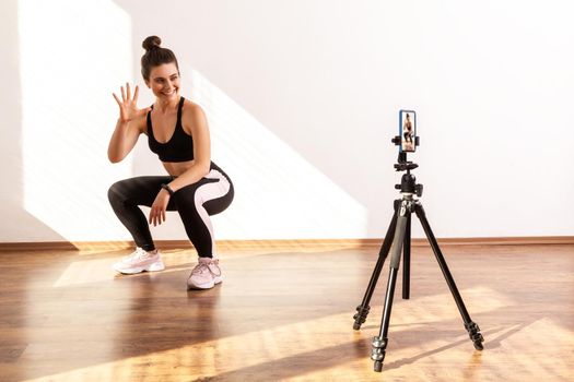 Coach recording video for her fitness vlog, squatting with followers, showing number of remaining, wearing black sports top and tights. Full length studio shot illuminated by sunlight from window.