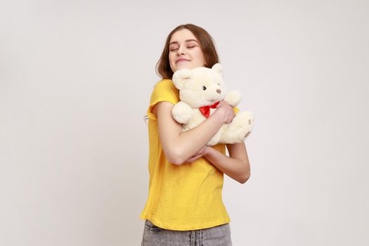 Satisfied happy teenager girl with brown hair in yellow casual T-shirt hugging cute white toy bear, closing eyes and dreaming, enjoying romantic gift. Indoor studio shot isolated on gray background.