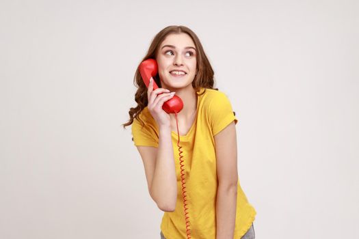 Positive happy young female with brown hair in yellow t-shirt talking landline telephone holding in hand handset, looking at away with dreamy look. Indoor studio shot isolated on gray background.