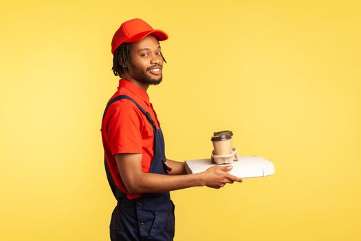 Side view portrait of optimistic professional courier standing with pizza box and coffee in disposable cup, fast food delivery service. Indoor studio shot isolated on yellow background.