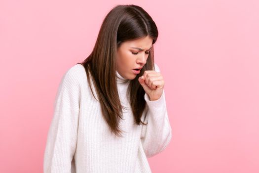 Profile portrait of young adult brunette sick woman coughing, catches cold, having high temperature, wearing white casual style sweater. Indoor studio shot isolated on pink background.