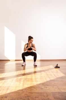 Sporty girl doing squatting sit up exercise while repeating after fitness coach online, using tablet, wearing black sports top and tights. Full length studio shot illuminated by sunlight from window.