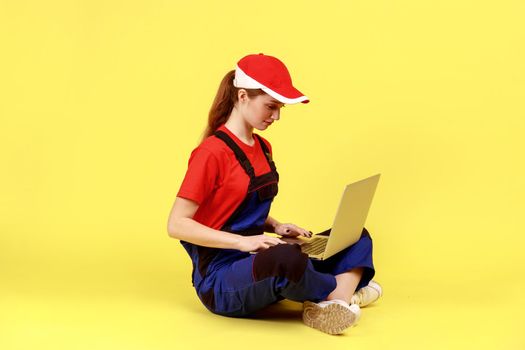 Side view portrait of concentrated handy woman sitting on floor and working on laptop, looking at display while typing, wearing overalls and red cap. Indoor studio shot isolated on yellow background.