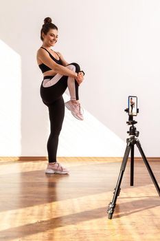 Slim fitness instructor recording online class of warming up before workout, stretching leg, wearing black sports top and tights. Full length studio shot illuminated by sunlight from window.