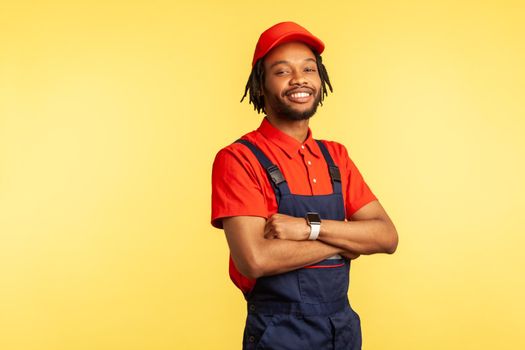 Satisfied handyman wearing overalls standing with crossed arms, looking at camera, profession of service industry, courier delivery. Indoor studio shot isolated on yellow background.