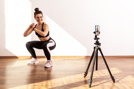 Smiling coach teaching correct squatting technique, recording tutorial video of workout, wearing black sports top and tights. Full length studio shot illuminated by sunlight from window.