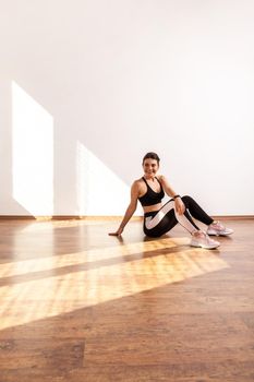 Side view of athletic female relaxing after sport, looking away, enjoying physical exercises at home, wearing black sports top and tights. Full length studio shot illuminated by sunlight from window.