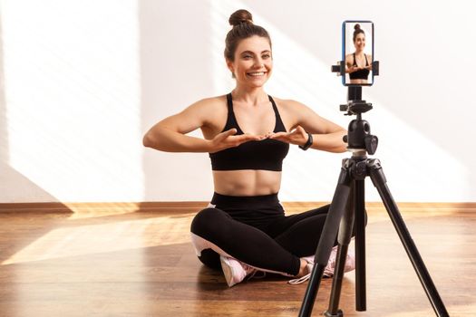 Female fitness instructor recording tutorial video of breathing technique after workout, wearing black sports top and tights. Full length studio shot illuminated by sunlight from window.