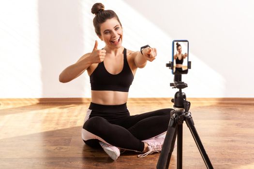 Happy fitness trainer pointing to phone camera and showing thumb up, praising you for online workout, wearing black sports top and tights. Full length studio shot illuminated by sunlight from window.