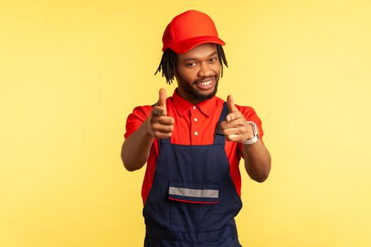 Portrait of positive bearded builder or courier wearing overalls, pointing to camera with fingers, looking with pleasant smile, service. Indoor studio shot isolated on yellow background.