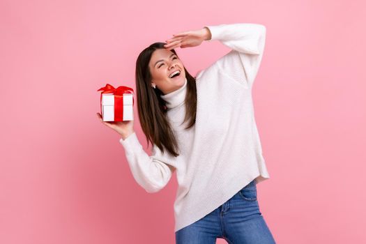 Portrait of satisfied female holding wrapped present box, looking far away with hand over forehead, wearing white casual style sweater. Indoor studio shot isolated on pink background.