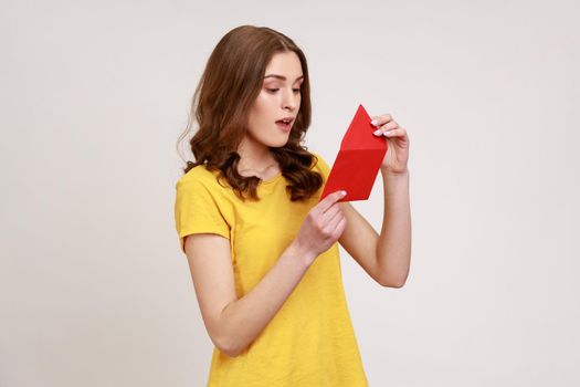 Side view of amazed young woman in yellow casual T-shirt opening envelope and reading letter, with surprised expression, got unexpected pleasant news. Indoor studio shot isolated on gray background.