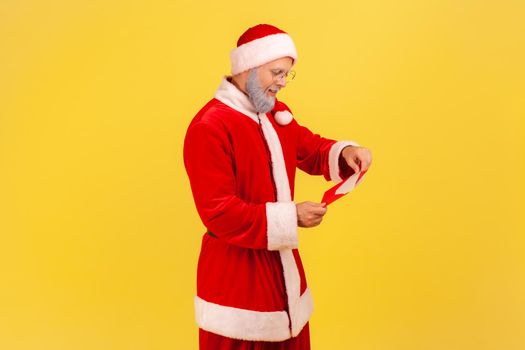 Side view of elderly man with gray beard in santa claus costume standing open red envelope with congratulations with winter holidays , reading letter. Indoor studio shot isolated on yellow background.