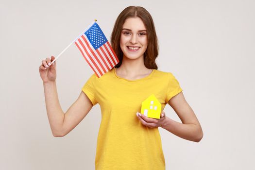 Portrait of happy teenager with brown hair holding flag of united states of america and paper house, dreaming about living in usa, green card. Indoor studio shot isolated on gray background.