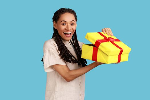 Portrait of happy amazed woman with black dreadlocks looking into gift box, opening present and looking at camera with happiness, wearing white shirt. Indoor studio shot isolated on blue background.