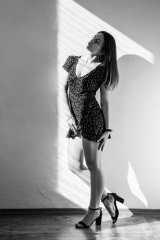 Beautiful sexy woman standing with closed eyes and dreamy facial expression, wearing dress and sandals. Black and white photography, indoor studio shot illuminated by sunlight from window.