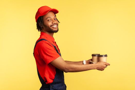 Side view portrait of smiling positive courier man wearing red T-shirt and blue uniform, giving take away coffee to client, delivery order. Indoor studio shot isolated on yellow background.