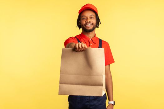Portrait of positive smiling deliveryman in overalls giving ordered parcel, delivering food in paper bag, post mail services. Indoor studio shot isolated on yellow background.