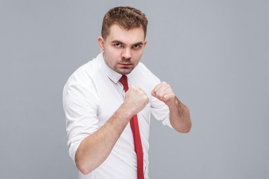 Portrait of young handsome serious man in white shirt and tie standing in boxing fists and looking at camera and ready to attack. indoor isolated on gray background.