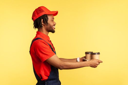 Delivery service. Side view, friendly deliveryman wearing blue overalls offering coffee, giving drinks in disposable cups and smiling. Indoor studio shot isolated on yellow background.