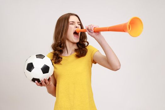 Extremely positive satisfied teenager girl in yellow t-shirt screaming in bullhorn holding soccer ball, celebrating victory of favorite football team. Indoor studio shot isolated on gray background.