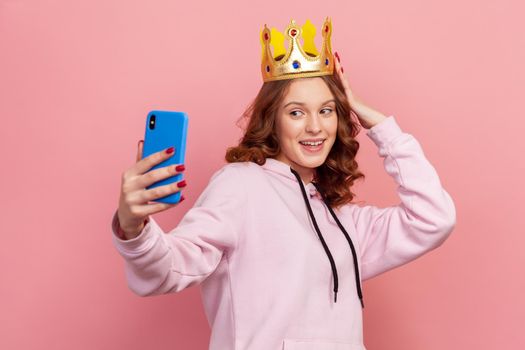 Portrait of smiling curly haired teenage girl in hoodie with gold crown posing on smartphone camera, taking selfie. Indoor studio shot isolated on pink background