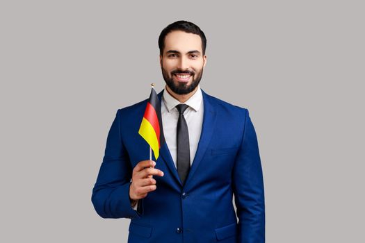 Delighted businessman holding Germany flag, celebrating Day of Germany - 3th October, expressing positive emotions, wearing official style suit. Indoor studio shot isolated on gray background.