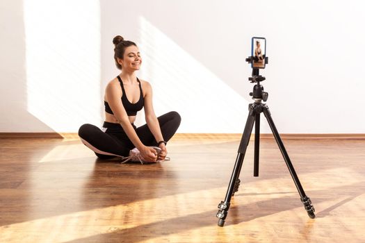 Female coach filming video of relaxing after workout, sitting on lotus pose with closed eyes, wearing black sports top and tights. Full length studio shot illuminated by sunlight from window.